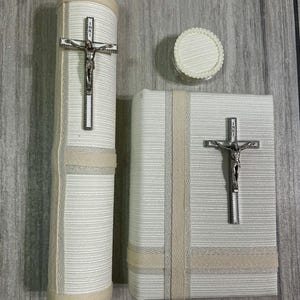 Puede incluir: Un conjunto religioso que presenta una vela cilíndrica alta, un pequeño recipiente redondo y un libro rectangular. Cada artículo está envuelto en una tela texturizada de color blanquecino con cintas beige. Una cruz plateada está fijada a la vela y al libro.