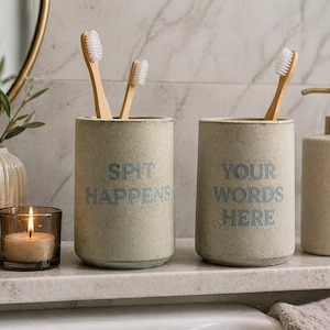 May include: A bathroom countertop scene features two cylindrical toothbrush holders with the phrases "SPIT HAPPENS" and "YOUR WORDS HERE" in blue. Two bamboo toothbrushes are in the holders. A soap dispenser and a lit candle are also present.