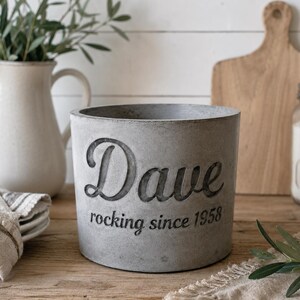Könnte beinhalten: Ein grauer Betonblumentopf mit dem Namen "Dave" und dem Schriftzug "rocking since 1958" auf der Seite. Der zylindrische Topf steht auf einer Holzoberfläche. Eine weiße Kanne und ein Schneidebrett befinden sich im Hintergrund.