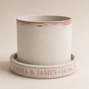 May include: A cylindrical light grey planter with a dusty rose-coloured brushstroke rim. It sits on a matching circular tray featuring the engraved text EMMA & JAMES and the date 14.06.25 in dusty rose lettering around the outer edge.