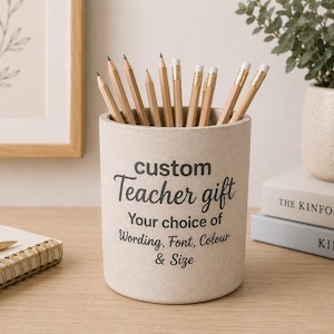 May include: A cylindrical beige pencil holder with a speckled texture, featuring black printed text that reads: custom Teacher gift Your choice of Wording, Font, Colour & Size. The holder is filled with several wooden pencils with erasers.