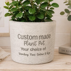Op de afbeelding: Een cilindervormige, gespikkelde lichtgrijze plantenpot met een getextureerde afwerking. De voorzijde bevat donkergrijze tekst die luidt: Custom made Plant Pot, Your choice of Wording Font Colour & Size. Een groene bladplant staat in de pot.