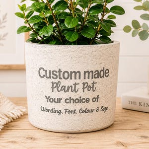 Op de afbeelding: Een cilindervormige, gespikkelde gebroken witte plantenpot met donkergrijze tekst: Custom made Plant Pot, Your choice of Wording, Font, Colour & Size. De pot bevat een groene plant en heeft een textuur die aanvoelt als steen.