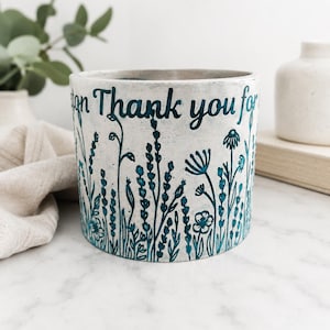 May include: A cylindrical white planter featuring a textured surface with dark teal botanical illustrations of wildflowers and grasses. The upper portion of the planter displays the printed text "Thank you for" in a dark teal, sans-serif font.