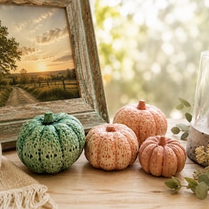 May include: Three small decorative pumpkins with textured surfaces, one in a mottled green finish and two in a speckled orange-brown tone, arranged near a rustic wooden picture frame containing a landscape photograph of a sunset over a field.