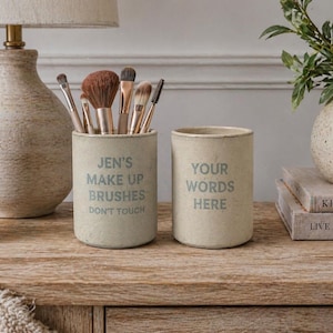 May include: Two cylindrical beige containers with text, one reading "JEN'S MAKE UP BRUSHES DON'T TOUCH" and the other "YOUR WORDS HERE." The first container holds makeup brushes. The containers sit on a wooden surface.