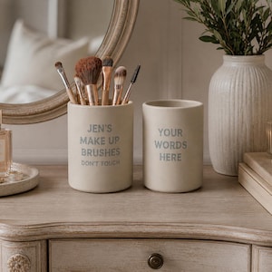 May include: Two off-white ceramic containers on a wooden vanity. One holds makeup brushes and reads "Jen's Make Up Brushes Don't Touch." The other reads "Your Words Here." A vase with greenery and a candle are also present.