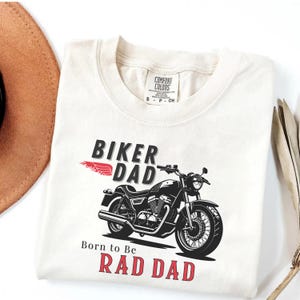 May include: Cream-colored t-shirt with a motorcycle graphic and the words "Biker Dad" and "Born to be Rad Dad" in black and red. The shirt has a crew neck and short sleeves.