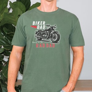 May include: Olive green t-shirt featuring a motorcycle graphic with the text "BIKER DAD" and "Born to Be RAD DAD". The design includes a red wing graphic. The shirt is a short-sleeved crew neck.