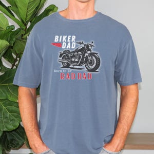 May include: Blue t-shirt with a motorcycle graphic and the words "BIKER DAD" and "RAD DAD". The motorcycle is black and silver, and the text is in white and red. The shirt is a solid color and has short sleeves.