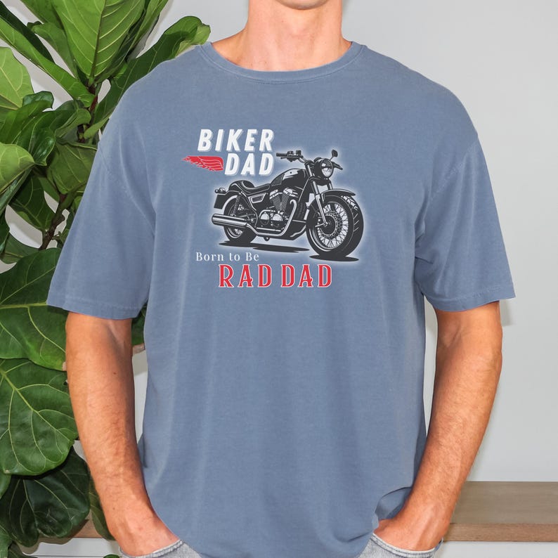 May include: Blue t-shirt with a motorcycle graphic and the words "BIKER DAD" and "RAD DAD". The motorcycle is black and silver, and the text is in white and red. The shirt is a solid color and has short sleeves.