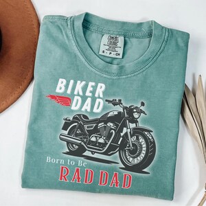 May include: A teal Comfort Colors t-shirt featuring a motorcycle graphic and the words "BIKER DAD" and "Born to Be RAD DAD" in white and red. The shirt has a crew neck and short sleeves.