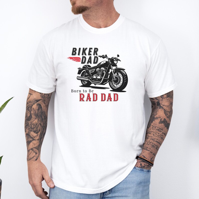 May include: White t-shirt featuring a black motorcycle graphic with the text "BIKER DAD" and "Born to Be RAD DAD" in black and red. The design includes a red wing element. The shirt is made of a soft material.
