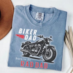 May include: A blue Comfort Colors t-shirt with the words "BIKER DAD" and "Born to Be RAD DAD" in white and red. A black motorcycle graphic is featured below the text. The shirt has a crew neck and short sleeves.