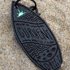 May include: Black wooden surfboard-shaped keychain with the words "FOLLOW THE CURRENT WHEREVER IT FLOWS" engraved. Features a small, light green design at the top and wave and eye details. The keychain is on a black ring.