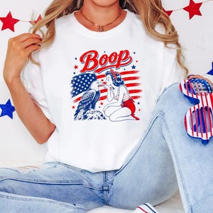 May include: A white crewneck sweatshirt featuring a graphic print of a pin-up style figure booping a bald eagle on the beak. The design includes the text Boop, a stylized American flag background, and red and blue stars. The shirt is paired with light blue denim jeans.