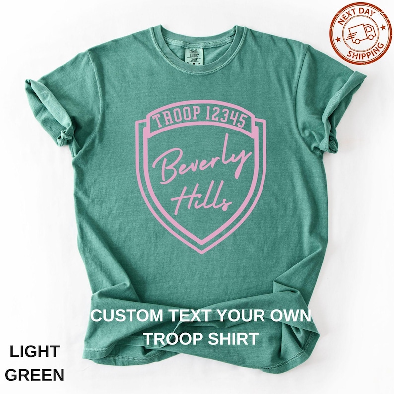 A green t-shirt with a pink shield-shaped logo that says "Troop 123445 Beverly Hills".