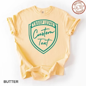 A yellow t-shirt with a green shield-shaped logo that says "Troop 12345 Custom Text" printed on it.