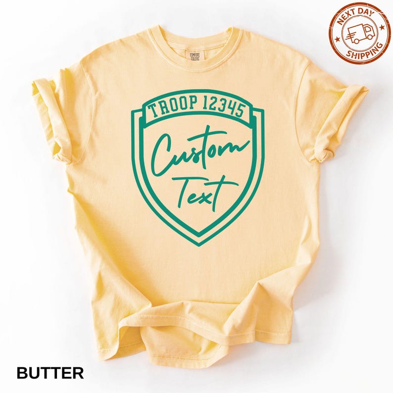 A yellow t-shirt with a green shield-shaped logo that says "Troop 12345 Custom Text" printed on it.