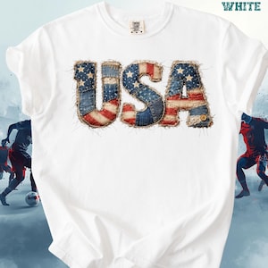 May include: White t-shirt with the word "USA" in a patchwork design, featuring the American flag's colors and stars. The shirt has a crew neck and short sleeves. The word "WHITE" is printed at the top right.