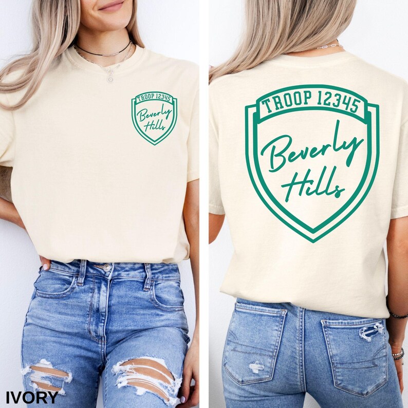 A woman wearing a cream-colored t-shirt with a green shield-shaped logo and text on the back, paired with distressed blue jeans.
