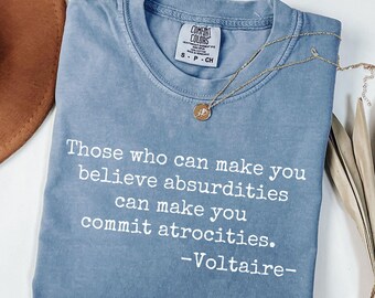 May include: A bluejean Comfort Colors t-shirt with the quote "Those who can make you believe absurdities can make you commit atrocities. -Voltaire-" printed in white. A gold necklace with a pendant is on the shirt.