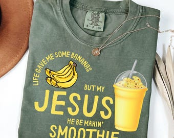 May include: Moss green t-shirt with the text "Life gave me some bananas but my Jesus he be makin' smoothie" in yellow. The shirt features a banana graphic and a yellow smoothie illustration. A silver necklace is draped across the shirt.