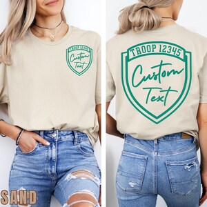 A woman wearing a beige t-shirt with a green shield-shaped logo on the back, and jeans.