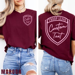 A woman wearing a burgundy t-shirt with a shield-shaped logo and text on the back, paired with distressed blue jeans.