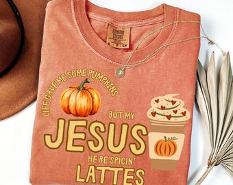 May include: A coral-colored t-shirt with the text "Life Gave Me Some Pumpkins But My Jesus He Be Spicin' Lattes." The shirt features a pumpkin graphic and a coffee cup with hearts. A gold necklace is draped across the shirt.