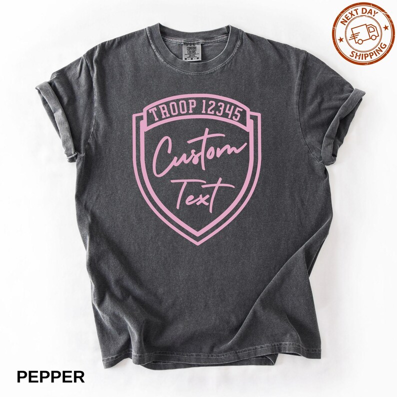 A gray t-shirt with a pink shield-shaped logo that says "Troop 12345 Custom Text" printed on it.