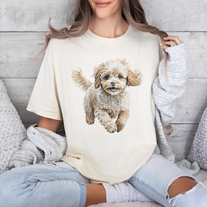 May include: A cream-colored short-sleeve t-shirt featuring a detailed, realistic illustration of a fluffy, light brown puppy with curly fur, floppy ears, and a happy expression, centered on the front of the garment.