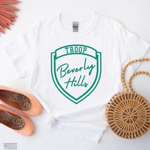 a white T-shirt with a green shield-shaped logo, a pair of peach-colored ballet flats, a woven straw bag, and a pair of white shoes.
