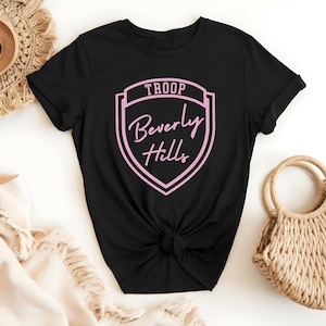 A black t-shirt with a pink shield-shaped logo that says "Troop Beverly Hills" printed on it, along with a beige woven purse and a straw hat.