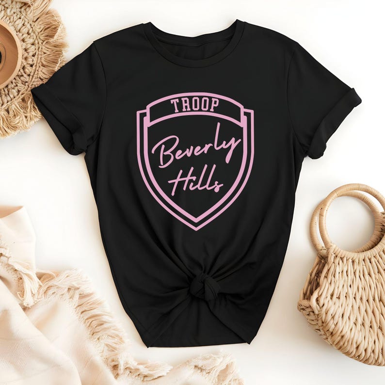 A black t-shirt with a pink shield-shaped logo that says "Troop Beverly Hills" printed on it, along with a beige woven purse and a straw hat.
