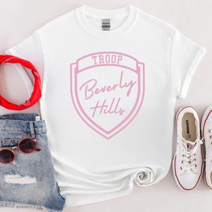 A white T-shirt with a pink shield design and the text "Troop Beverly Hills" printed on it, along with a pair of denim shorts, a red bandana, sunglasses, and a pair of white sneakers.