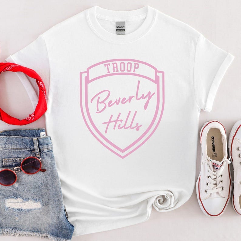 A white T-shirt with a pink shield design and the text "Troop Beverly Hills" printed on it, along with a pair of denim shorts, a red bandana, sunglasses, and a pair of white sneakers.