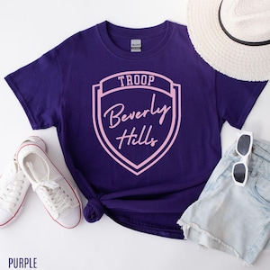 Purple T-shirt with a shield design, sunglasses, a hat, and a pair of white sneakers.