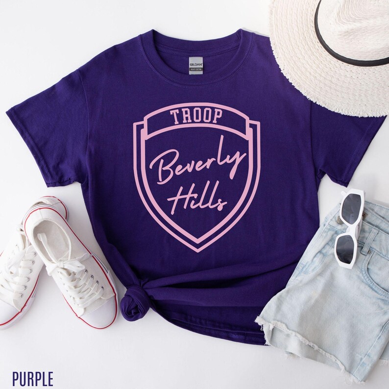 Purple T-shirt with a shield design, sunglasses, a hat, and a pair of white sneakers.