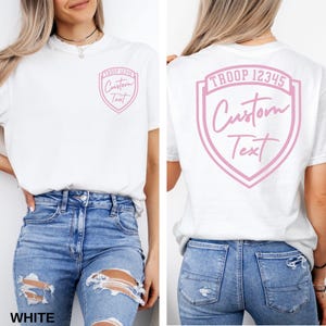 A woman wearing a white t-shirt with a pink shield design and text on the back.