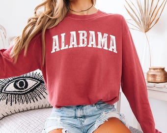 May include: A crimson long-sleeve crewneck sweatshirt featuring the word ALABAMA printed in a white, distressed, arched font across the chest. The garment has a relaxed fit with ribbed cuffs and a ribbed neckline, showcasing a vintage-inspired look.