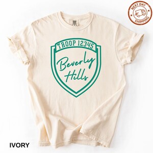 A cream-colored T-shirt with a green shield-shaped logo that says "Troop 12345 Beverly Hills".
