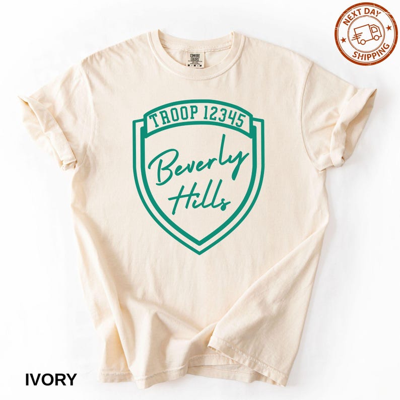 A cream-colored T-shirt with a green shield-shaped logo that says "Troop 12345 Beverly Hills".