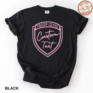 A black t-shirt with a pink shield-shaped logo that says "Troop 12345 Custom Text" printed on it.