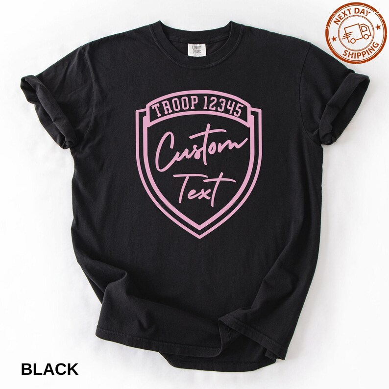 A black t-shirt with a pink shield-shaped logo that says "Troop 12345 Custom Text" printed on it.