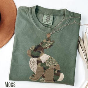 Pode incluir: Uma camiseta verde musgo com um design de coelho de retalhos com padrões de camuflagem e florais. O coelho tem um olho de botão e um detalhe de renda. Um colar de ouro com um pingente está drapeado na camisa.