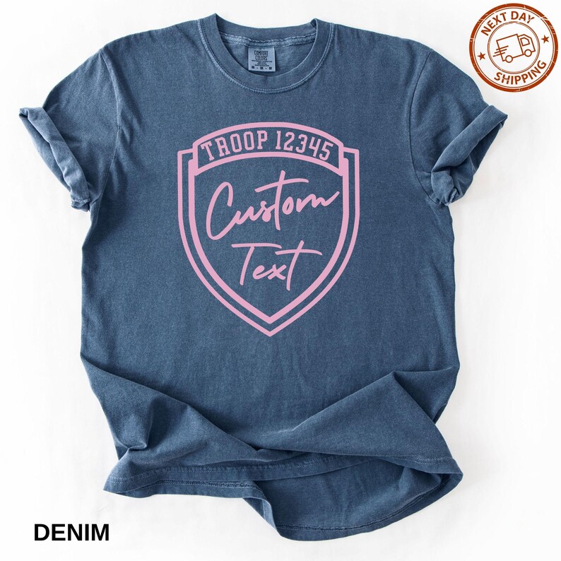 A navy blue t-shirt with a pink shield-shaped logo that says "Troop 12345 Custom Text" printed on it.