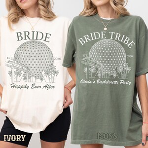 Puede incluir: Una camiseta color marfil con un gráfico verde oscuro de un domo geodésico, monorraíl y palmeras, con el texto BRIDE, EST. 2026 y Happily Ever After. Una camiseta verde musgo con un gráfico blanco del mismo domo, el texto BRIDE TRIBE, EST. 2026 y Olivia's Bachelorette Party.