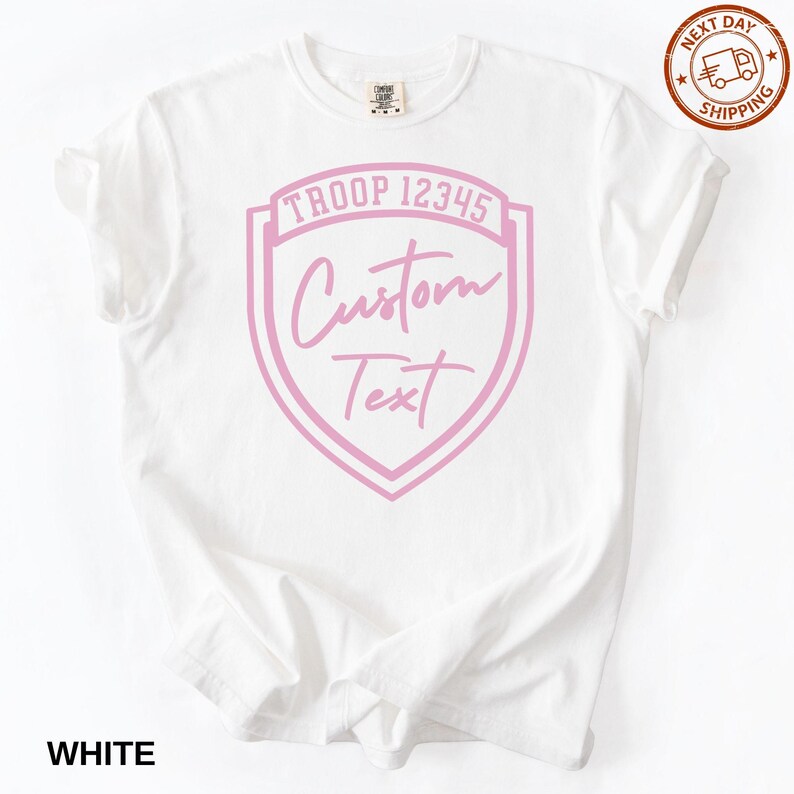 A white T-shirt with a pink shield-shaped logo that says "Troop 12345 Custom Text" printed on it.