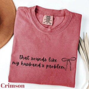 Könnte beinhalten: Karmesinrotes T-Shirt mit dem Text "that sounds like my husband's problem" und einer Schleifengrafik. Das Shirt ist von der Marke Comfort Colors, Größe S-P-CH. Das Shirt liegt auf einer weißen Oberfläche.